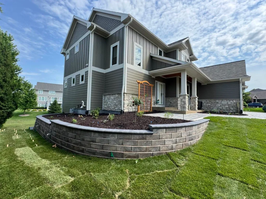 Curved block retaining wall around a raised planting bed beside a gray house
