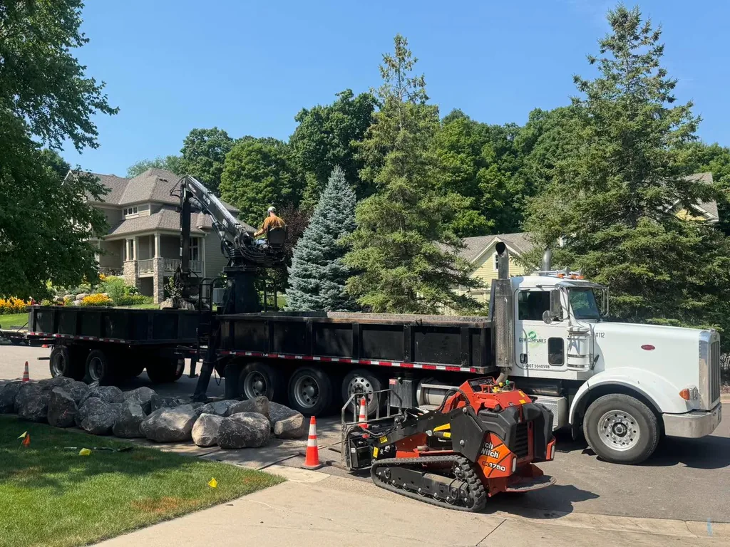 Dump truck and skid-steer delivering landscape material beside a home