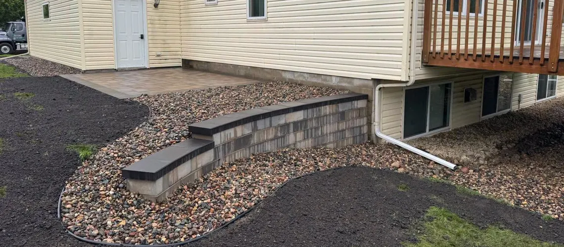 Curved block retaining wall with dark capstones holding a decorative rock bed beside a home